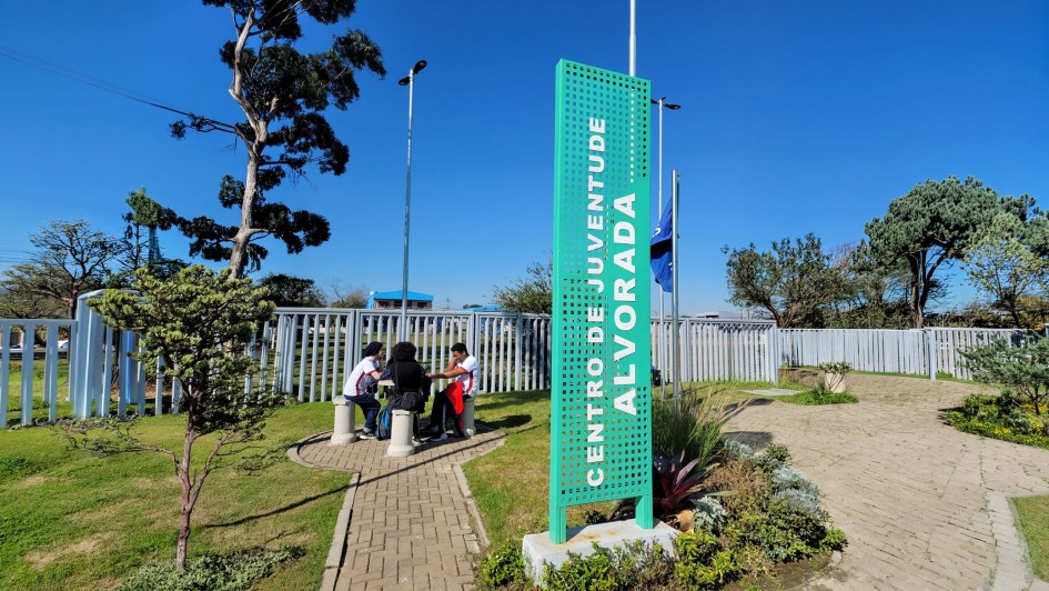 Foto mostra entrada do CJ Alvorada com placa em primeiro plano e jardim ao fundo, onde est&atilde;o tr&ecirc;s jovens sentados em volta de uma mesa circular no centro do jardim. O c&eacute;u &eacute; azul e h&aacute; &aacute;rvores no fundo do jardim. 