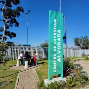Foto mostra entrada do CJ Alvorada com placa em primeiro plano e jardim ao fundo, onde est&atilde;o tr&ecirc;s jovens sentados em volta de uma mesa circular no centro do jardim. O c&eacute;u &eacute; azul e h&aacute; &aacute;rvores no fundo do jardim. 