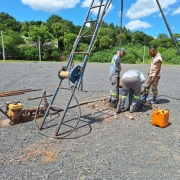 Trabalhadores realizam a&ccedil;&otilde;es de medi&ccedil;&atilde;o no terreno com britas. Ao fundo, um bosque verde e o c&eacute;u azul. 