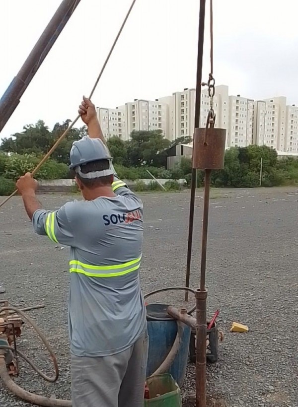 Trabalhador mexe em um andaime no terreno de brita. Ao fundo, um conjunto de prédios. 