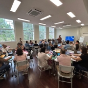 Foto mostra sala com coordenadores regionais em mesas circulares. Eles acompanham instru&ccedil;&otilde;es do palestrante. A sala tem janelas grandes que mostram &aacute;rvores. 