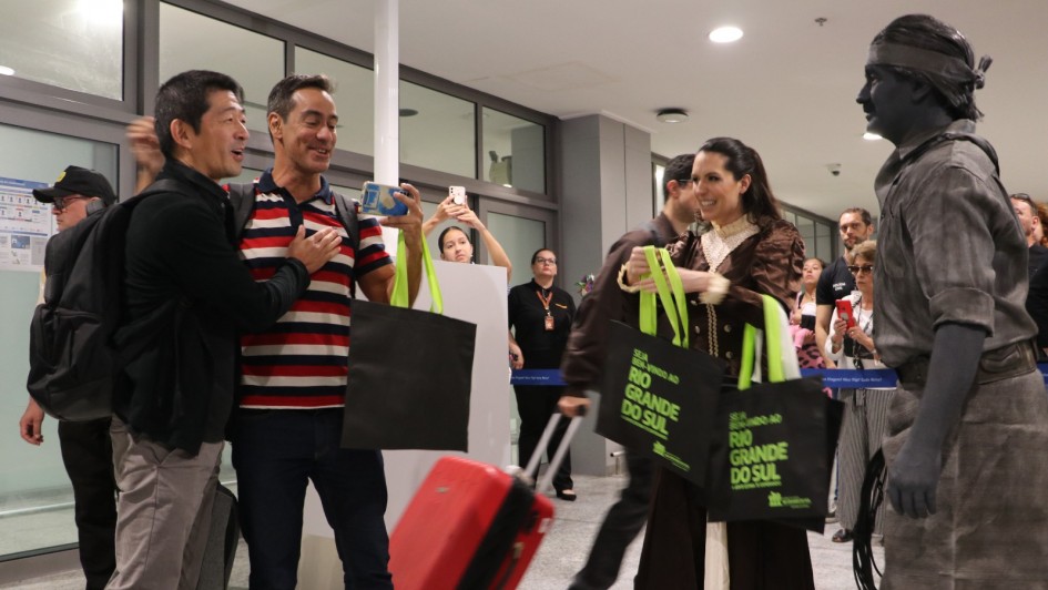 Recep&ccedil;&atilde;o Tur&iacute;stica no Rio Grande do Sul
A imagem retrata um momento de recep&ccedil;&atilde;o calorosa em um ambiente interno moderno e bem iluminado, possivelmente o sagu&atilde;o de um aeroporto ou centro de eventos.

Intera&ccedil;&atilde;o &agrave; Esquerda: Dois homens aparecem em primeiro plano, sorrindo e se cumprimentando. Um deles veste uma jaqueta preta e o outro uma camisa polo listrada em vermelho, azul e branco. Este &uacute;ltimo segura um smartphone e uma sacola preta com al&ccedil;as verdes.

Figura Central (Cultura Local): No centro da imagem, uma mulher vestida com um traje t&iacute;pico ga&uacute;cho (vestido longo marrom escuro com detalhes em renda e gola clara) segura duas sacolas promocionais pretas. As sacolas possuem al&ccedil;as verdes e a frase "SEJA BEM-VINDO AO RIO GRANDE DO SUL" estampada em letras verdes vibrantes.

Est&aacute;tua Viva: No canto direito, um artista perform&aacute;tico atua como uma est&aacute;tua viva. Ele est&aacute; inteiramente coberto por tinta cinza-chumbo, incluindo pele e roupas (camisa r&uacute;stica, cal&ccedil;as e uma faixa na cabe&ccedil;a), mantendo uma pose est&aacute;tica enquanto segura o que parece ser um chicote ou corda.

Cen&aacute;rio e Detalhes: O ambiente possui piso de granito claro, teto branco com ilumina&ccedil;&atilde;o embutida e grandes divis&oacute;rias de vidro ao fundo. Uma mala de viagem vermelha com rodas est&aacute; posicionada no ch&atilde;o entre os personagens principais, refor&ccedil;ando o contexto de viagem e turismo.