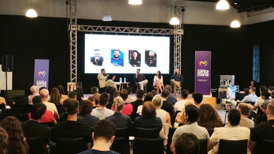 Foto mostra ao fundo quatro palestrantes sentados, um deles fala no microfone. A plateia, de costas, acompanha a apresenta&ccedil;&atilde;o. No fundo do palco, h&aacute; um tel&atilde;o com a foto dos palestrantes.  