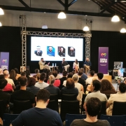 Foto mostra ao fundo quatro palestrantes sentados, um deles fala no microfone. A plateia, de costas, acompanha a apresenta&ccedil;&atilde;o. No fundo do palco, h&aacute; um tel&atilde;o com a foto dos palestrantes.  