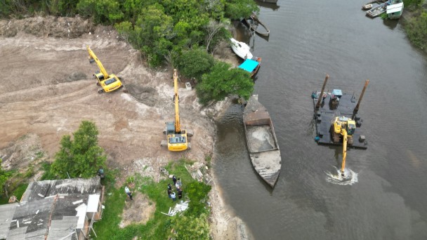 Foto a&eacute;rea mostra rio com barco e draga em atividade e na margem com duas por&ccedil;&otilde;es de mata e no centro um terreno j&aacute; limpo por duas m&aacute;quinas retroescavadeiras. 