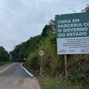 A imagem mostra um obra de pavimenta&ccedil;&atilde;o asf&aacute;ltica. No lado direito da foto, h&aacute; uma placa onde se l&ecirc; "Obra em parceria com o governo do Estado". 