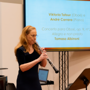 A musicista Viktoria Tatour toca oboé em uma sala iluminada, posicionada diante de um telão que exibe informações de um concerto, incluindo os nomes dos intérpretes e o repertório. Ela está ao lado de um piano de cauda e segura o instrumento com postura concentrada, enquanto o ambiente sugere um momento de apresentação ou ensaio.
