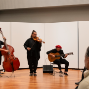Três músicos se apresentam na Sala de Recitais da Ospa, formando um trio com contrabaixo, violino e violão. Eles tocam posicionados em semicírculo, com instrumentos amplificados e caixas de som próximas, enquanto um pequeno público assiste sentado.