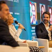 Em um palco de evento, tr&ecirc;s pessoas est&atilde;o sentadas em poltronas brancas participando de um painel. Leite, de terno escuro, segura um microfone e sorri enquanto olha para uma mulher ao lado, que tamb&eacute;m fala ao microfone. Ao fundo, um tel&atilde;o azul exibe fotos dos participantes e textos do evento.