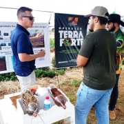 A fotografia retrata uma demonstra&ccedil;&atilde;o t&eacute;cnica durante um evento ao ar livre, identificado pelo nome do arquivo como a ExpoAgro Cotricampo. A cena foca na dissemina&ccedil;&atilde;o de conhecimento sobre manejo e conserva&ccedil;&atilde;o de recursos naturais.

Contexto e Pessoas
A&ccedil;&atilde;o Principal: Um homem &agrave; esquerda, vestindo uma camisa polo azul e &oacute;culos de sol, gesticula enquanto explica conte&uacute;dos t&eacute;cnicos para dois visitantes.

Visitantes: &Agrave; direita, dois homens escutam a explica&ccedil;&atilde;o. Um deles usa uma camiseta verde e bon&eacute; cinza; o outro veste uma camisa com estampas que remetem &agrave; bandeira do Brasil e um chap&eacute;u de aba larga preto.

Elementos de Demonstra&ccedil;&atilde;o
Cartazes T&eacute;cnicos: Atr&aacute;s do palestrante, h&aacute; um cartaz branco com o t&iacute;tulo "Manejo, propriedades f&iacute;sicas [do] solo e da &aacute;gua", contendo diagramas e fotos explicativas. Ao lado, um banner vertical preto exibe o logotipo da "Opera&ccedil;&atilde;o Terra Forte".

Experimento Pr&aacute;tico: Em primeiro plano, sobre uma bancada branca, h&aacute; uma demonstra&ccedil;&atilde;o did&aacute;tica utilizando garrafas pl&aacute;sticas cortadas. As garrafas cont&ecirc;m diferentes camadas de solo e cobertura vegetal (palha), provavelmente utilizadas para mostrar visualmente processos de infiltra&ccedil;&atilde;o de &aacute;gua e prote&ccedil;&atilde;o do solo contra eros&atilde;o.

Ambiente
Cen&aacute;rio: O evento ocorre em uma &aacute;rea aberta com gramado e canteiros de plantas ao fundo. Estruturas de metal e lonas sugerem a montagem de estandes t&iacute;picos de feiras agr&iacute;colas.