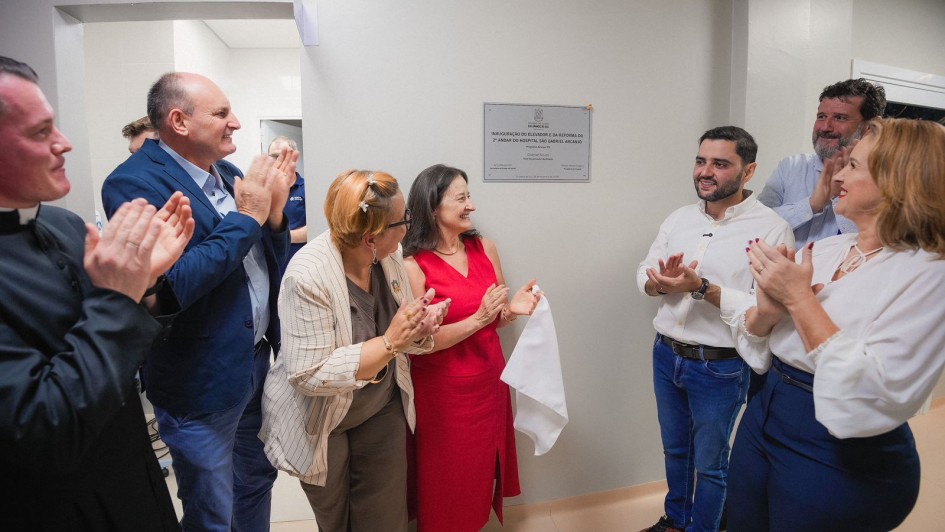 A imagem mostra um grupo de pessoas em p&eacute;, batendo palmas e sorrindo, em frente a uma placa de inaugura&ccedil;&atilde;o afixada na parede branca de um hospital. 