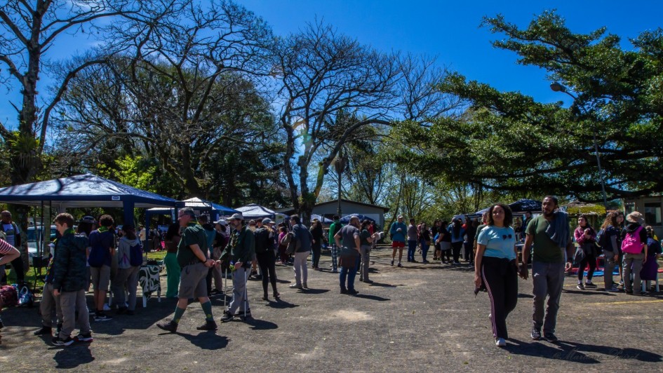 Foto mostra um espa&ccedil;o aberto com cal&ccedil;amento e cercado de &aacute;rvores. H&aacute; uma feira com barracas e p&uacute;blico circulando. O c&eacute;u ao fundo &eacute; azul, sem nuvens. 