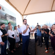 Eduardo Leite fala ao microfone em um palanque sob uma tenda branca. Ele veste camisa branca e cal&ccedil;a azul. Ao seu lado, autoridades acompanham o discurso em frente a um painel com a logomarca do Governo do Rio Grande do Sul e proje&ccedil;&otilde;es da futura escola. &Agrave; direita, fot&oacute;grafos e cinegrafistas registram o evento.