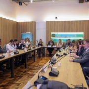 Esta &eacute; uma descri&ccedil;&atilde;o detalhada da imagem para fins de acessibilidade:

A imagem registra uma reuni&atilde;o formal ocorrida em uma sala ampla e bem iluminada. Cerca de vinte pessoas, entre homens e mulheres com trajes executivos (ternos, camisas e blusas sociais), est&atilde;o sentadas ao redor de uma grande mesa de madeira clara em formato de "U".

Detalhes do Ambiente
Mobili&aacute;rio: A mesa principal ocupa grande parte do espa&ccedil;o. &Agrave; frente de cada participante, h&aacute; microfones pretos individuais de mesa, copos de papel brancos e, em alguns pontos, notebooks e documentos. As cadeiras s&atilde;o de escrit&oacute;rio, na cor preta.

Estrutura da Sala: As paredes possuem pain&eacute;is de madeira clara que v&atilde;o at&eacute; meia altura, com a parte superior em branco. O piso &eacute; de madeira em estilo parquet (tacos). No fundo da sala, h&aacute; uma estante embutida com ilumina&ccedil;&atilde;o suave, exibindo pequenas bandeiras e objetos decorativos.

Tecnologia: Na parede &agrave; esquerda, um monitor de TV exibe um documento com o cronograma ou pauta da reuni&atilde;o.

Composi&ccedil;&atilde;o da Cena
Os participantes est&atilde;o distribu&iacute;dos pelas laterais e pela cabeceira da mesa (ao fundo da imagem). Alguns est&atilde;o focados em quem fala, outros consultam celulares ou fazem anota&ccedil;&otilde;es. O clima &eacute; de uma reuni&atilde;o de trabalho institucional e s&eacute;ria. &Agrave; esquerda, em destaque, v&ecirc;-se um banner vertical azul e branco.