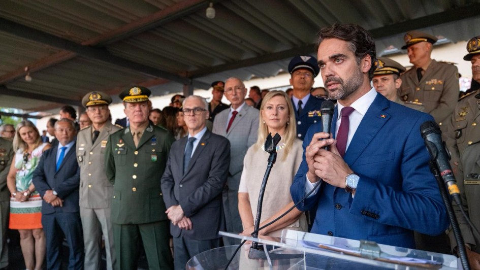 A imagem registra um evento oficial em que o Governador do Rio Grande do Sul, Eduardo Leite, discursa para uma plateia de autoridades civis e militares.

Composi&ccedil;&atilde;o da Imagem
Primeiro Plano: &Agrave; direita, Eduardo Leite aparece em destaque, falando ao microfone em um p&uacute;lpito de acr&iacute;lico transparente. Ele tem cabelos e barba aparados, veste um palet&oacute; azul marinho, camisa branca e gravata bord&ocirc;. Ele segura o microfone com as duas m&atilde;os e olha para o lado oposto &agrave; c&acirc;mera.

Segundo Plano: Logo atr&aacute;s do governador, diversas autoridades acompanham o discurso. Est&atilde;o presentes homens e mulheres em trajes formais (ternos e vestidos) e v&aacute;rios oficiais militares de alta patente, vestindo fardas em tons de verde-oliva e azul, adornadas com ins&iacute;gnias e medalhas.

Ambiente: A cerim&ocirc;nia ocorre em um local aberto, mas sob uma cobertura de estrutura met&aacute;lica aparente. A ilumina&ccedil;&atilde;o &eacute; natural, sugerindo um evento realizado durante o dia em um p&aacute;tio ou pavilh&atilde;o.

Clima e Tom
A cena transmite uma atmosfera de solenidade e formalidade, t&iacute;pica de cerim&ocirc;nias de posse e an&uacute;ncios governamentais. As express&otilde;es dos presentes s&atilde;o s&eacute;rias e atentas.