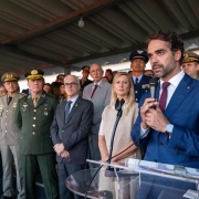 A imagem registra um evento oficial em que o Governador do Rio Grande do Sul, Eduardo Leite, discursa para uma plateia de autoridades civis e militares.

Composi&ccedil;&atilde;o da Imagem
Primeiro Plano: &Agrave; direita, Eduardo Leite aparece em destaque, falando ao microfone em um p&uacute;lpito de acr&iacute;lico transparente. Ele tem cabelos e barba aparados, veste um palet&oacute; azul marinho, camisa branca e gravata bord&ocirc;. Ele segura o microfone com as duas m&atilde;os e olha para o lado oposto &agrave; c&acirc;mera.

Segundo Plano: Logo atr&aacute;s do governador, diversas autoridades acompanham o discurso. Est&atilde;o presentes homens e mulheres em trajes formais (ternos e vestidos) e v&aacute;rios oficiais militares de alta patente, vestindo fardas em tons de verde-oliva e azul, adornadas com ins&iacute;gnias e medalhas.

Ambiente: A cerim&ocirc;nia ocorre em um local aberto, mas sob uma cobertura de estrutura met&aacute;lica aparente. A ilumina&ccedil;&atilde;o &eacute; natural, sugerindo um evento realizado durante o dia em um p&aacute;tio ou pavilh&atilde;o.

Clima e Tom
A cena transmite uma atmosfera de solenidade e formalidade, t&iacute;pica de cerim&ocirc;nias de posse e an&uacute;ncios governamentais. As express&otilde;es dos presentes s&atilde;o s&eacute;rias e atentas.