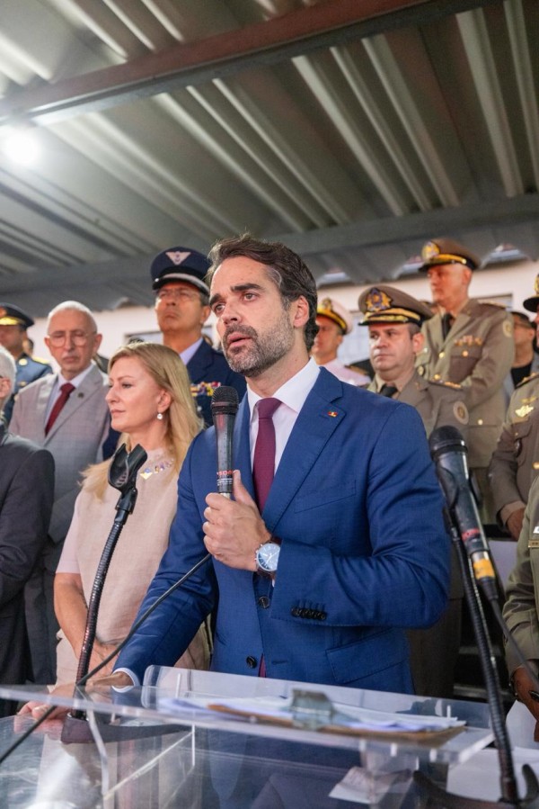 As imagens retratam uma cerimônia oficial de posse e anúncio de investimentos no Rio Grande do Sul. Abaixo, as descrições detalhadas para fins de acessibilidade:

Imagem 1: Posse do Comando-Geral da Brigada Militar
Esta imagem apresenta um plano médio lateral do governador Eduardo Leite durante seu discurso.

O Governador: Localizado à direita da foto, Eduardo Leite está posicionado atrás de um púlpito de acrílico transparente. Ele veste um paletó azul, camisa branca e gravata bordô, segurando o microfone com as duas mãos enquanto fala.

Autoridades e Convidados: Atrás do governador, uma fileira de autoridades acompanha o ato. Entre elas, destacam-se diversos oficiais militares em uniformes formais (verde-oliva, caqui e azul-escuro) com medalhas e quepe. Há também civis em trajes sociais, incluindo uma mulher de blusa bege e um homem de terno cinza com gravata vermelha.

Cenário: O evento ocorre sob uma estrutura de teto metálico aparente, com iluminação natural.

Imagem 2: Destaque aos Investimentos em Estrutura
Esta imagem é um plano mais fechado (close-up) da mesma cena anterior, focando na expressão do governador.

Expressão: Eduardo Leite é mostrado em um ângulo ligeiramente inferior, olhando para frente com uma expressão séria e compenetrada enquanto discursa.

Segundo Plano: A profundidade de campo é menor, deixando as autoridades ao fundo um pouco menos nítidas, mas ainda identificáveis, reforçando o foco na fala do governador sobre os investimentos realizados.

Detalhes Técnicos: O microfone preto e a borda do púlpito de acrílico aparecem na parte inferior da moldura.