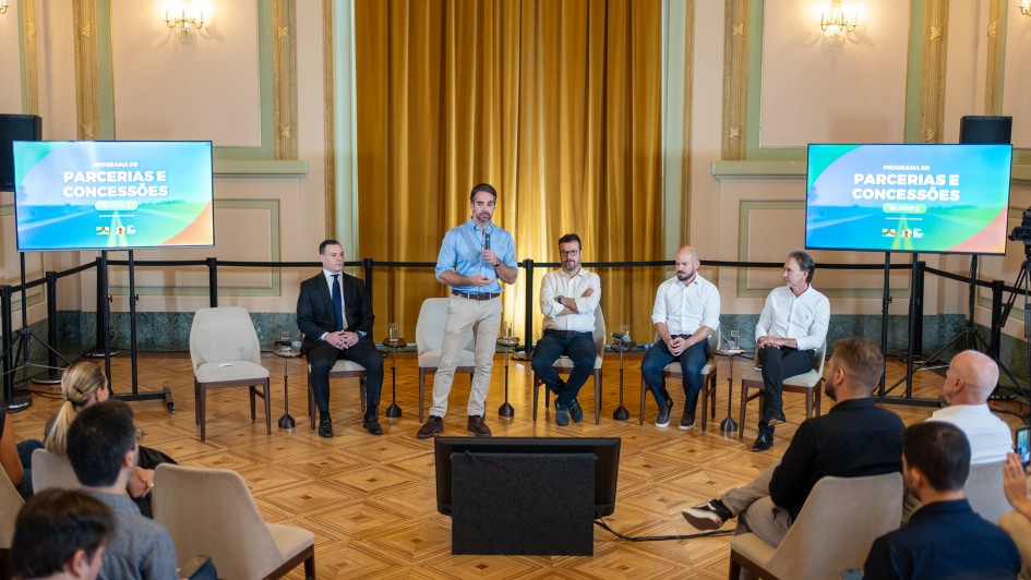 Sal&atilde;o amplo com piso de madeira e cortinas douradas ao fundo. Cinco homens est&atilde;o &agrave; frente: um em p&eacute; falando ao microfone e quatro sentados em cadeiras ao lado. Duas telas exibem &ldquo;Programa de Parcerias e Concess&otilde;es &ndash; Bloco 2&rdquo;. H&aacute; p&uacute;blico sentado de frente para eles.