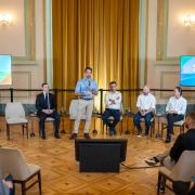 Sal&atilde;o amplo com piso de madeira e cortinas douradas ao fundo. Cinco homens est&atilde;o &agrave; frente: um em p&eacute; falando ao microfone e quatro sentados em cadeiras ao lado. Duas telas exibem &ldquo;Programa de Parcerias e Concess&otilde;es &ndash; Bloco 2&rdquo;. H&aacute; p&uacute;blico sentado de frente para eles.