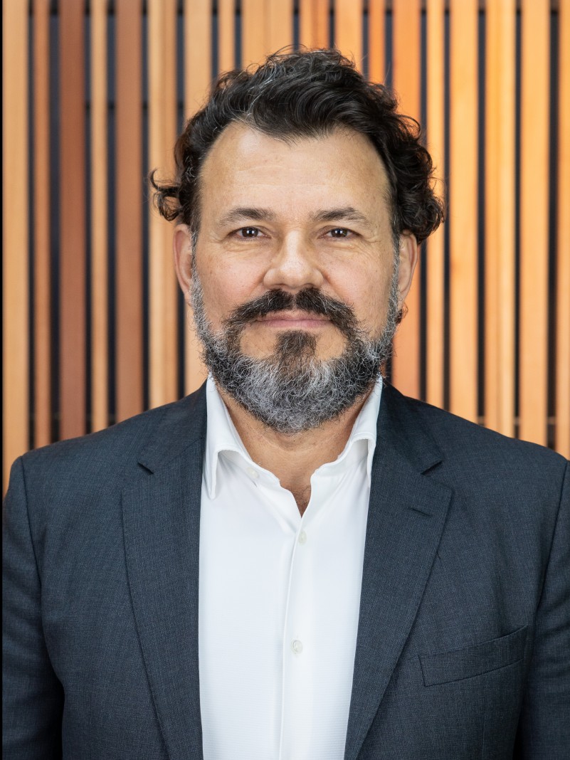 Foto posada em primeiro plano de Antonio, homem de cabelo preto curto e barba grisalha e que usa terno. Ele sorri olhando para a c&acirc;mera. O fundo &eacute; de um material que parece madeira clara.