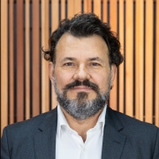 Foto posada em primeiro plano de Antonio, homem de cabelo preto curto e barba grisalha e que usa terno. Ele sorri olhando para a c&acirc;mera. O fundo &eacute; de um material que parece madeira clara.