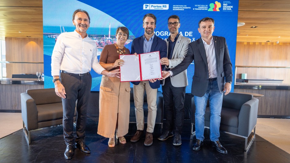 A imagem registra o momento da assinatura de um protocolo de inten&ccedil;&otilde;es para um investimento de R$ 130 milh&otilde;es no Distrito Industrial do Rio Grande.

Composi&ccedil;&atilde;o e Participantes
O Governador: No centro da foto, o governador Eduardo Leite segura uma pasta vermelha aberta contendo o documento assinado. Ele veste um blazer azul-marinho, camisa social azul-clara e cal&ccedil;a bege.

Autoridades: Ao lado do governador, quatro outras autoridades (tr&ecirc;s homens e uma mulher) tamb&eacute;m seguram as bordas da pasta, simbolizando a coopera&ccedil;&atilde;o entre as partes. Eles vestem trajes casuais-sociais, como blazers sem gravata e camisas claras.

Cen&aacute;rio: O grupo est&aacute; posicionado sobre um tablado preto em uma sala de arquitetura moderna, com forro e pain&eacute;is de madeira.

Elementos Visuais de Fundo
Painel Digital: Atr&aacute;s do grupo, um grande tel&atilde;o azul exibe as logomarcas da Portos RS, da Secretaria de Desenvolvimento Econ&ocirc;mico, da Secretaria de Log&iacute;stica e Transportes e o bras&atilde;o do Governo do Estado do Rio Grande do Sul.

Identidade Visual: O tel&atilde;o tamb&eacute;m apresenta uma fotografia a&eacute;rea do complexo portu&aacute;rio e industrial, refor&ccedil;ando o tema do investimento anunciado.