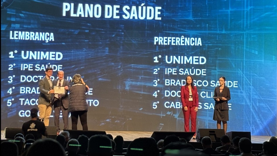 Foto mostra duas pessoas no palco recebendo o certificado com a lista de planos de sa&uacute;de citados na pesquisa listados no tel&atilde;o ao fundo.