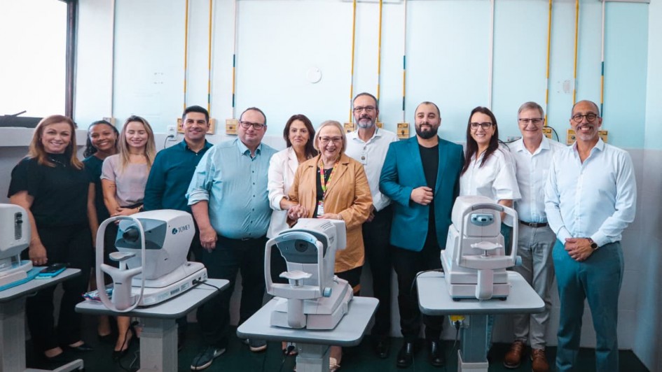 A fotografia retrata um grupo de pessoas em um ambiente hospitalar, posando atr&aacute;s de novos equipamentos m&eacute;dicos especializados.

Elementos Principais
Equipamentos Oftalmol&oacute;gicos: Em primeiro plano, destacam-se tr&ecirc;s aparelhos oftalmol&oacute;gicos modernos de cor branca, montados sobre suportes cinzas ajust&aacute;veis com rodas. No aparelho da esquerda, &eacute; poss&iacute;vel ler a marca "TOMEY".

O Grupo: Onze pessoas (seis homens e cinco mulheres) est&atilde;o perfiladas lado a lado atr&aacute;s dos equipamentos. Elas vestem trajes que variam entre o casual-chique e o formal. No centro, destaca-se uma mulher de &oacute;culos, cabelos loiros e blazer caramelo, que sorri para a c&acirc;mera.

Segundo Plano e Contexto
Ambiente Cl&iacute;nico: O grupo est&aacute; em uma sala de exames com paredes azul-claro. Na parte superior da parede, h&aacute; uma rede de tubula&ccedil;&otilde;es amarelas expostas e v&aacute;rias tomadas el&eacute;tricas, elementos t&iacute;picos de instala&ccedil;&otilde;es de sa&uacute;de.

Atmosfera: A imagem possui uma ilumina&ccedil;&atilde;o clara e direta, sugerindo um evento de inaugura&ccedil;&atilde;o ou entrega oficial de equipamentos.