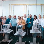 A fotografia retrata um grupo de pessoas em um ambiente hospitalar, posando atr&aacute;s de novos equipamentos m&eacute;dicos especializados.

Elementos Principais
Equipamentos Oftalmol&oacute;gicos: Em primeiro plano, destacam-se tr&ecirc;s aparelhos oftalmol&oacute;gicos modernos de cor branca, montados sobre suportes cinzas ajust&aacute;veis com rodas. No aparelho da esquerda, &eacute; poss&iacute;vel ler a marca "TOMEY".

O Grupo: Onze pessoas (seis homens e cinco mulheres) est&atilde;o perfiladas lado a lado atr&aacute;s dos equipamentos. Elas vestem trajes que variam entre o casual-chique e o formal. No centro, destaca-se uma mulher de &oacute;culos, cabelos loiros e blazer caramelo, que sorri para a c&acirc;mera.

Segundo Plano e Contexto
Ambiente Cl&iacute;nico: O grupo est&aacute; em uma sala de exames com paredes azul-claro. Na parte superior da parede, h&aacute; uma rede de tubula&ccedil;&otilde;es amarelas expostas e v&aacute;rias tomadas el&eacute;tricas, elementos t&iacute;picos de instala&ccedil;&otilde;es de sa&uacute;de.

Atmosfera: A imagem possui uma ilumina&ccedil;&atilde;o clara e direta, sugerindo um evento de inaugura&ccedil;&atilde;o ou entrega oficial de equipamentos.