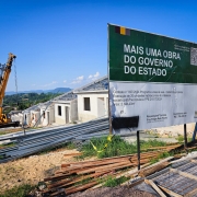Esta &eacute; uma descri&ccedil;&atilde;o detalhada da imagem para fins de acessibilidade:

Descri&ccedil;&atilde;o da Imagem
A fotografia registra um canteiro de obras de um conjunto habitacional em est&aacute;gio intermedi&aacute;rio de constru&ccedil;&atilde;o, sob um c&eacute;u azul sem nuvens.

Placa Informativa (Primeiro Plano): No lado direito da imagem, h&aacute; uma grande placa de metal com as cores do estado (verde, vermelho e amarelo no topo).

O texto em destaque diz: "MAIS UMA OBRA DO GOVERNO DO ESTADO".

Informa&ccedil;&otilde;es t&eacute;cnicas detalham o Contrato n&ordm; 007/2024 para o Programa A Casa &eacute; Sua - Calamidade, no munic&iacute;pio de Estrela.

A placa menciona a execu&ccedil;&atilde;o de 20 unidades habitacionais de interesse social, com um investimento total de R$ 2.785.400,00.

As Constru&ccedil;&otilde;es: Ao fundo, observa-se uma fileira de casas geminadas em constru&ccedil;&atilde;o, dispostas em um terreno com leve declive. As paredes externas est&atilde;o em reboco cinza e os telhados apresentam estruturas met&aacute;licas de suporte, ainda sem as telhas.

Maquin&aacute;rio e Materiais:

&Agrave; esquerda, um guindaste amarelo de grande porte est&aacute; posicionado na rua lateral, com o bra&ccedil;o estendido em dire&ccedil;&atilde;o &agrave;s casas.

No centro, h&aacute; pilhas de perfis met&aacute;licos e vigas no ch&atilde;o, prontos para uso.

&Agrave; direita, no canto inferior, v&ecirc;-se uma estrutura de vergalh&otilde;es de ferro sobre o solo, preparada para a concretagem de uma cal&ccedil;ada ou funda&ccedil;&atilde;o.

Paisagem: O horizonte mostra uma &aacute;rea verde com colinas e vegeta&ccedil;&atilde;o densa, sob uma luz solar intensa que projeta sombras n&iacute;tidas no terreno.