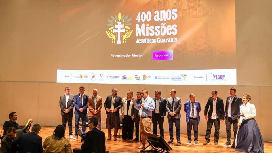 Foto mostra autoridades no palco em fileira. No alto, uma tela mostra uma proje&ccedil;&atilde;o com a logo do evento dos 400 anos das Miss&otilde;es Jesu&iacute;ticas Guaranis.
