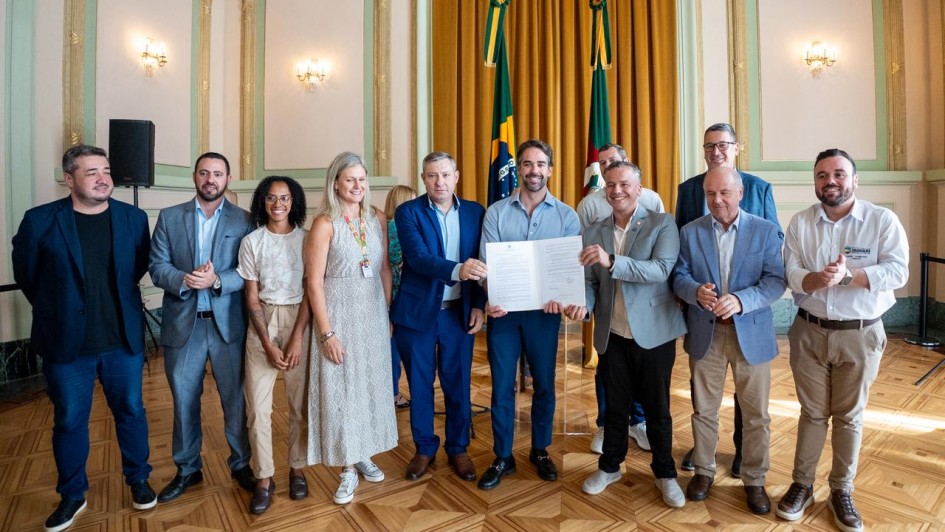 Esta &eacute; uma descri&ccedil;&atilde;o detalhada da imagem para fins de acessibilidade:

Descri&ccedil;&atilde;o da Imagem
A fotografia registra o ato oficial de lan&ccedil;amento do programa Passe F&aacute;cil Estudantil, realizado em um sal&atilde;o solene com arquitetura cl&aacute;ssica.

A&ccedil;&atilde;o Central: No centro da imagem, o Governador Eduardo Leite (vestindo camisa social cinza azulada e cal&ccedil;a escura) e outros dois homens seguram juntos um documento oficial aberto com duas p&aacute;ginas, exibindo-o para a c&acirc;mera.

O Grupo: Ao redor deles, outras sete pessoas (cinco homens e duas mulheres) posam para a foto em uma linha horizontal. O grupo veste trajes que variam entre o social e o casual-executivo. Uma das mulheres &agrave; esquerda utiliza um crach&aacute; oficial com cord&atilde;o colorido.

O Ambiente:

O grupo est&aacute; sobre um piso de madeira (parquet) com padr&otilde;es geom&eacute;tricos claros e escuros.

Ao fundo, h&aacute; um grande painel de cortinas douradas que emoldura as bandeiras do Brasil e do Rio Grande do Sul, posicionadas em mastros centrais.

As paredes s&atilde;o de tom creme com detalhes em relevo verde claro e arandelas cl&aacute;ssicas acesas.

&Agrave; esquerda, nota-se uma caixa de som sobre um pedestal, indicando a realiza&ccedil;&atilde;o de uma cerim&ocirc;nia com sistema de &aacute;udio.

Contexto: Conforme o t&iacute;tulo do arquivo, o evento marca uma iniciativa para ampliar o acesso &agrave; educa&ccedil;&atilde;o no estado atrav&eacute;s do Passe F&aacute;cil Estudantil.