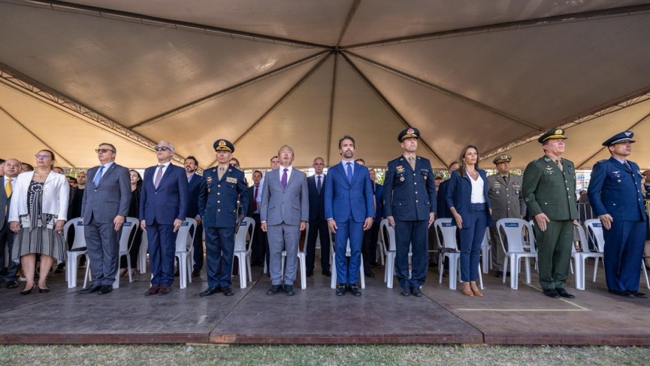A foto mostra uma fileira de autoridades e integrantes do Corpo de Bombeiros posicionados em p&eacute; sobre um palco elevado. Algumas pessoas vestem uniformes militares escuros ou verdes, enquanto outras usam trajes sociais formais. 