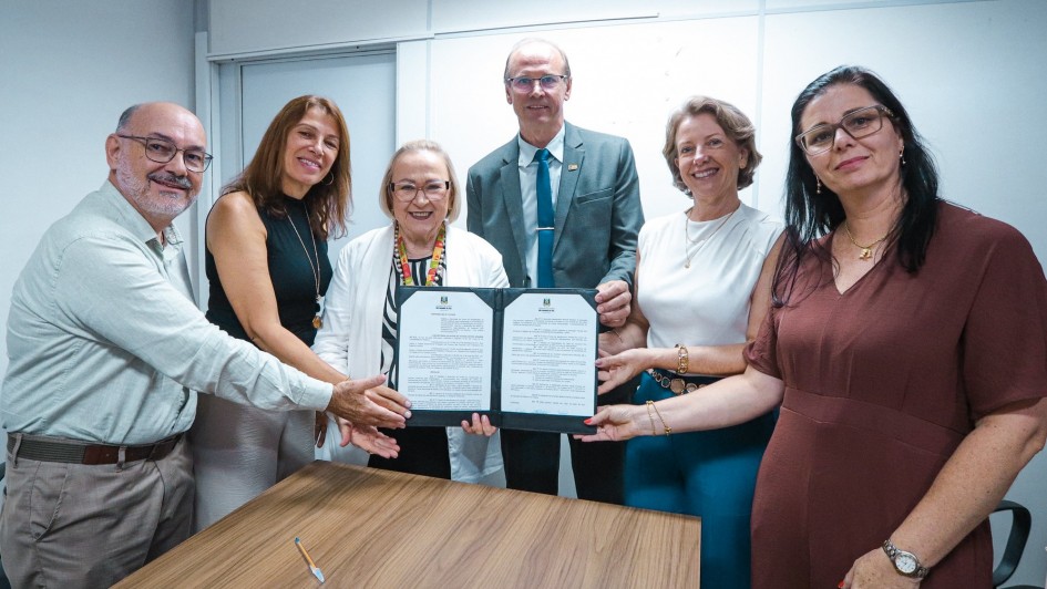 Esta &eacute; uma descri&ccedil;&atilde;o detalhada da imagem para fins de acessibilidade:

Descri&ccedil;&atilde;o da Imagem
A fotografia registra o ato formal de assinatura de um conv&ecirc;nio em um ambiente de escrit&oacute;rio com ilumina&ccedil;&atilde;o clara e paredes brancas.

A&ccedil;&atilde;o Central: Um grupo de seis pessoas (quatro mulheres e dois homens) est&aacute; posicionado atr&aacute;s de uma mesa de madeira clara. Eles seguram juntos uma pasta oficial preta aberta, exibindo um documento de duas p&aacute;ginas com o bras&atilde;o do Estado do Rio Grande do Sul no topo.

Os Participantes:

&Agrave; esquerda, um homem de camisa social clara e &oacute;culos sorri para a c&acirc;mera.

Ao lado dele, uma mulher de blusa preta sem mangas.

No centro, uma mulher de cabelos loiros curtos, usando &oacute;culos e um crach&aacute; oficial com cord&atilde;o colorido, segura a pasta.

Atr&aacute;s dela, um homem de terno cinza e gravata azul tamb&eacute;m segura o documento.

&Agrave; direita, uma mulher de blusa branca e outra de vestido marrom completam o grupo. Todos sorriem, transmitindo um clima de realiza&ccedil;&atilde;o.

Detalhes do Ambiente:

Sobre a mesa, em frente ao grupo, h&aacute; apenas uma caneta azul, indicando que a assinatura acabou de ocorrer.

O fundo &eacute; simples, composto por divis&oacute;rias de escrit&oacute;rio brancas, o que mant&eacute;m o foco total nas pessoas e no documento.

Contexto: De acordo com o t&iacute;tulo do arquivo, a imagem celebra a habilita&ccedil;&atilde;o do munic&iacute;pio de Torres ao programa TEAcolhe, com validade para o ano de 2026.
