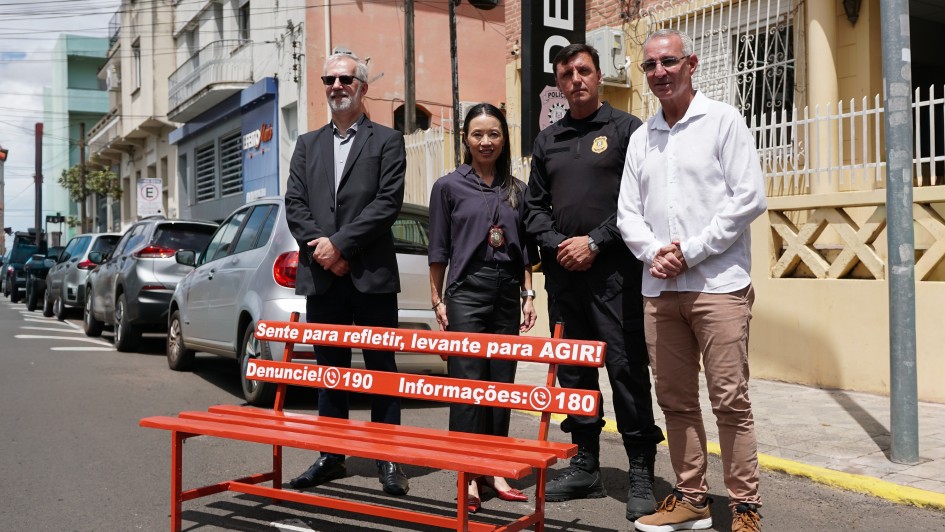 A imagem mostra um banco vermelho instalado em uma rua asfaltada, em &aacute;rea urbana. O banco &eacute; comprido, com estrutura fina e frases pintadas em branco no encosto, incluindo: &ldquo;Sente para refletir, levante para agir!&rdquo;, al&eacute;m de n&uacute;meros de contato: &ldquo;Denuncie: 190&rdquo; e &ldquo;Informa&ccedil;&otilde;es: 180&rdquo;. Atr&aacute;s do banco, quatro pessoas est&atilde;o posicionadas lado a lado, vestindo trajes sociais e uniformes institucionais.