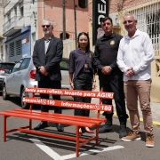 A imagem mostra um banco vermelho instalado em uma rua asfaltada, em &aacute;rea urbana. O banco &eacute; comprido, com estrutura fina e frases pintadas em branco no encosto, incluindo: &ldquo;Sente para refletir, levante para agir!&rdquo;, al&eacute;m de n&uacute;meros de contato: &ldquo;Denuncie: 190&rdquo; e &ldquo;Informa&ccedil;&otilde;es: 180&rdquo;. Atr&aacute;s do banco, quatro pessoas est&atilde;o posicionadas lado a lado, vestindo trajes sociais e uniformes institucionais.