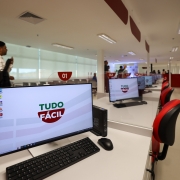 Foto de uma unidade do Tudo F&aacute;cil, de paredes e piso branco, com foco numa mesa de atendimento com computador. H&aacute; v&aacute;rias outras alinhadas atr&aacute;s. H&aacute; pessoas andando pelo local.