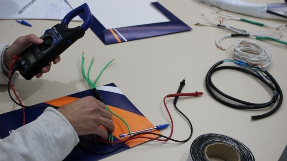 Foto em plano detalhe das m&atilde;os de um socioeducando mexendo com um aparelho eletr&ocirc;nico, com os bra&ccedil;os apoiados numa mesa clara. Sobre ela est&atilde;o fios e papeis. 