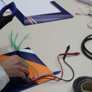 Foto em plano detalhe das m&atilde;os de um socioeducando mexendo com um aparelho eletr&ocirc;nico, com os bra&ccedil;os apoiados numa mesa clara. Sobre ela est&atilde;o fios e papeis. 