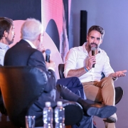 Tr&ecirc;s homens sentados em poltronas participam de uma conversa em um palco iluminado; um deles fala ao microfone enquanto os outros escutam atentamente.