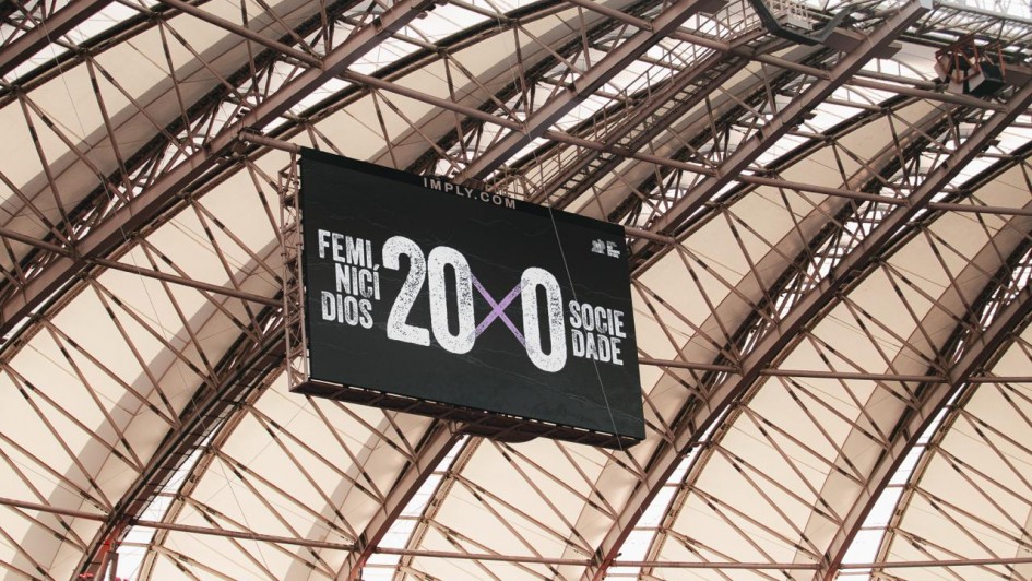 Um tel&atilde;o pendurado em uma estrutura met&aacute;lica de est&aacute;dio exibe a mensagem &ldquo;Feminic&iacute;dios 20 x 0 Sociedade&rdquo;, destacando um placar simb&oacute;lico contra a viol&ecirc;ncia de g&ecirc;nero.