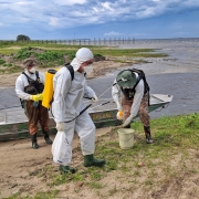 Foto mostra tr&ecirc;s servidores com roupas brancas, botas e m&aacute;scaras, preparando produtos para aplica&ccedil;&atilde;o. Eles est&atilde;o na beira da lagoa, pr&oacute;ximo a um barco. Ao fundo, a lagoa e o c&eacute;u carregado para chuva. 