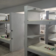 A foto mostra um ambiente interno com fileiras de estruturas cinzas que funcionam como beliches ou alojamentos modulares, cada uma com prateleiras embutidas. Sobre as superf&iacute;cies est&atilde;o organizados itens pessoais, como produtos de higiene e cobertores.