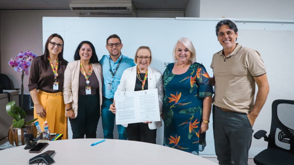 A fotografia mostra um grupo de seis pessoas posando para uma foto oficial em um ambiente de escrit&oacute;rio. Elas est&atilde;o alinhadas lado a lado, sorrindo, atr&aacute;s de uma mesa redonda de cor clara.

Elementos Centrais
O Documento: No centro do grupo, uma mulher de &oacute;culos e blazer branco segura um documento oficial aberto com as duas m&atilde;os, exibindo o texto e as assinaturas.

Os Integrantes (da esquerda para a direita):

Uma mulher de cabelos longos e escuros, usando blusa marrom e cal&ccedil;a amarela vibrante.

Uma mulher sorridente com blazer bege sobre blusa marrom.

Um homem de &oacute;culos e camisa social azul clara.

A mulher central de blazer branco mencionada anteriormente.

Uma mulher de cabelos loiros curtos, vestindo um vestido azul-marinho com estampas florais tropicais em tons de laranja e verde.

Um homem alto, de cabelos grisalhos, vestindo uma camisa polo bege e cal&ccedil;a cinza.

Detalhes e Ambiente
Identifica&ccedil;&atilde;o: Tr&ecirc;s das pessoas &agrave; esquerda utilizam crach&aacute;s com cord&otilde;es coloridos (com o padr&atilde;o de quebra-cabe&ccedil;a, s&iacute;mbolo da conscientiza&ccedil;&atilde;o do autismo).

Cen&aacute;rio: O fundo &eacute; composto por uma divis&oacute;ria branca simples. &Agrave; esquerda, h&aacute; um vaso com uma orqu&iacute;dea lil&aacute;s. Na mesa em primeiro plano, v&ecirc;-se uma caneta azul e um dispositivo de confer&ecirc;ncia telef&ocirc;nica (triangular).

Ilumina&ccedil;&atilde;o: A luz &eacute; frontal e uniforme, t&iacute;pica de ambientes internos de trabalho, destacando as express&otilde;es amig&aacute;veis do grupo.