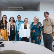 A fotografia mostra um grupo de seis pessoas posando para uma foto oficial em um ambiente de escrit&oacute;rio. Elas est&atilde;o alinhadas lado a lado, sorrindo, atr&aacute;s de uma mesa redonda de cor clara.

Elementos Centrais
O Documento: No centro do grupo, uma mulher de &oacute;culos e blazer branco segura um documento oficial aberto com as duas m&atilde;os, exibindo o texto e as assinaturas.

Os Integrantes (da esquerda para a direita):

Uma mulher de cabelos longos e escuros, usando blusa marrom e cal&ccedil;a amarela vibrante.

Uma mulher sorridente com blazer bege sobre blusa marrom.

Um homem de &oacute;culos e camisa social azul clara.

A mulher central de blazer branco mencionada anteriormente.

Uma mulher de cabelos loiros curtos, vestindo um vestido azul-marinho com estampas florais tropicais em tons de laranja e verde.

Um homem alto, de cabelos grisalhos, vestindo uma camisa polo bege e cal&ccedil;a cinza.

Detalhes e Ambiente
Identifica&ccedil;&atilde;o: Tr&ecirc;s das pessoas &agrave; esquerda utilizam crach&aacute;s com cord&otilde;es coloridos (com o padr&atilde;o de quebra-cabe&ccedil;a, s&iacute;mbolo da conscientiza&ccedil;&atilde;o do autismo).

Cen&aacute;rio: O fundo &eacute; composto por uma divis&oacute;ria branca simples. &Agrave; esquerda, h&aacute; um vaso com uma orqu&iacute;dea lil&aacute;s. Na mesa em primeiro plano, v&ecirc;-se uma caneta azul e um dispositivo de confer&ecirc;ncia telef&ocirc;nica (triangular).

Ilumina&ccedil;&atilde;o: A luz &eacute; frontal e uniforme, t&iacute;pica de ambientes internos de trabalho, destacando as express&otilde;es amig&aacute;veis do grupo.