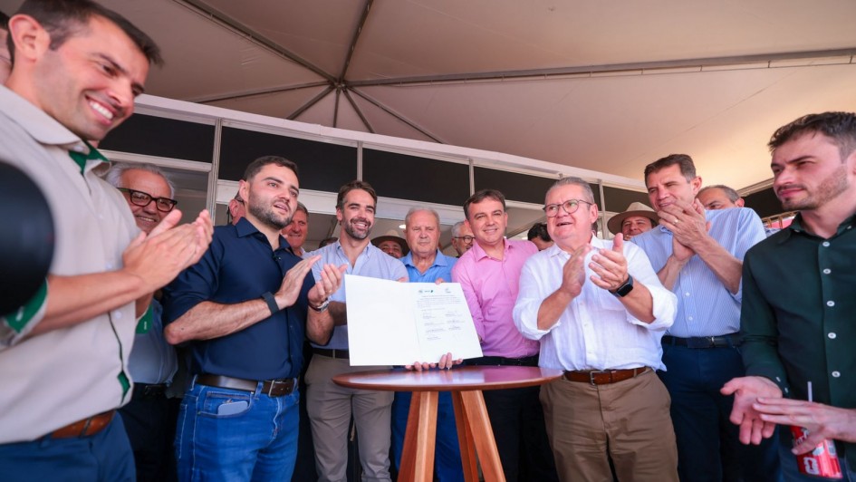 Esta &eacute; uma descri&ccedil;&atilde;o da imagem para fins de acessibilidade:

A fotografia registra um momento de celebra&ccedil;&atilde;o oficial durante a Expodireto Cotrijal, em que um grupo de autoridades e representantes do setor agropecu&aacute;rio se re&uacute;ne para a assinatura de documentos de investimento.

Elementos Principais
O Ato Central: Ao centro da imagem, sobre uma mesa alta e redonda de madeira (estilo bistr&ocirc;), um documento oficial aberto &eacute; exibido para a c&acirc;mera. V&aacute;rias m&atilde;os o seguram levemente pelas bordas.

As Autoridades (da esquerda para a direita):

Em destaque &agrave; esquerda, o vice-governador Gabriel Souza, vestindo uma camisa social azul-marinho e cal&ccedil;a jeans, aplaude com um sorriso.

Logo atr&aacute;s dele, ligeiramente ao centro, o governador Eduardo Leite, vestindo uma camisa azul clara de mangas curtas, observa o documento com uma express&atilde;o sorridente.

&Agrave; direita da mesa, um homem de camisa branca e cal&ccedil;a c&aacute;qui aplaude com entusiasmo.

Diversos outros homens em trajes casuais e sociais comp&otilde;em o grupo ao fundo, todos sorrindo ou aplaudindo, refor&ccedil;ando o clima de conquista.

Detalhes e Ambiente
Cen&aacute;rio: O evento ocorre sob uma grande tenda branca, com estruturas met&aacute;licas vis&iacute;veis no teto. Ao fundo, divis&oacute;rias de stands indicam que o local &eacute; um pavilh&atilde;o de feiras.

Ilumina&ccedil;&atilde;o e Composi&ccedil;&atilde;o: A foto foi tirada de um &acirc;ngulo ligeiramente de baixo para cima (contra-picado), o que confere uma sensa&ccedil;&atilde;o de import&acirc;ncia ao momento. A luz &eacute; clara e bem distribu&iacute;da, t&iacute;pica de eventos diurnos em ambientes cobertos.

Clima: A atmosfera &eacute; de otimismo e coopera&ccedil;&atilde;o institucional, evidenciada pela linguagem corporal de celebra&ccedil;&atilde;o (palmas e sorrisos) de todos os presentes.