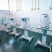 Sala de exames oftalmol&oacute;gicos com quatro equipamentos especializados alinhados lado a lado sobre suportes com rod&iacute;zios. A sala tem piso escuro, paredes claras e pontos de energia organizados na parede.