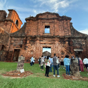 A imagem mostra um grupo espalhado pelo gramado observando atentamente a imponente fachada de pedra das ruínas missioneiras. A construção histórica, marcada por tons de vermelho escuro e detalhes arquitetônicos preservados, ocupa quase toda a largura da imagem. O céu azul com nuvens finas completa a cena.
