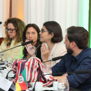 A imagem mostra participantes sentados ao redor de uma mesa de reunião com microfones direcionados para eles. Há copos com água, blocos e bandeiras sobre a mesa. Uma pessoa gesticula enquanto conversa com outra ao lado. O fundo é formado por cortinas claras iluminadas por luzes coloridas que criam um efeito suave no ambiente.
