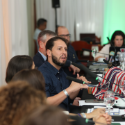 A imagem mostra participantes sentados lado a lado em uma mesa longa de reunião, conversando enquanto utilizam microfones e observam documentos e telas à frente. Há bandeiras pequenas sobre a mesa, além de copos de água e anotações. O ambiente é iluminado por luzes coloridas projetadas nas cortinas ao fundo.
